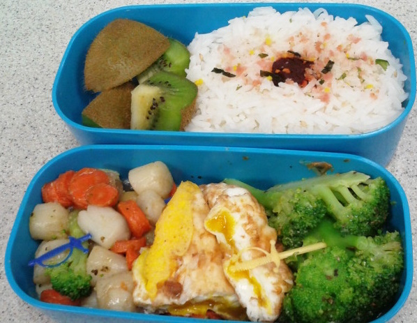 blue bento box showing rice with pickled plum and shrimp furikake and kiwi chunks in one half, and pan cooked scallops carrots and broccoli with rolled omlette chunks with sword shaped food picks